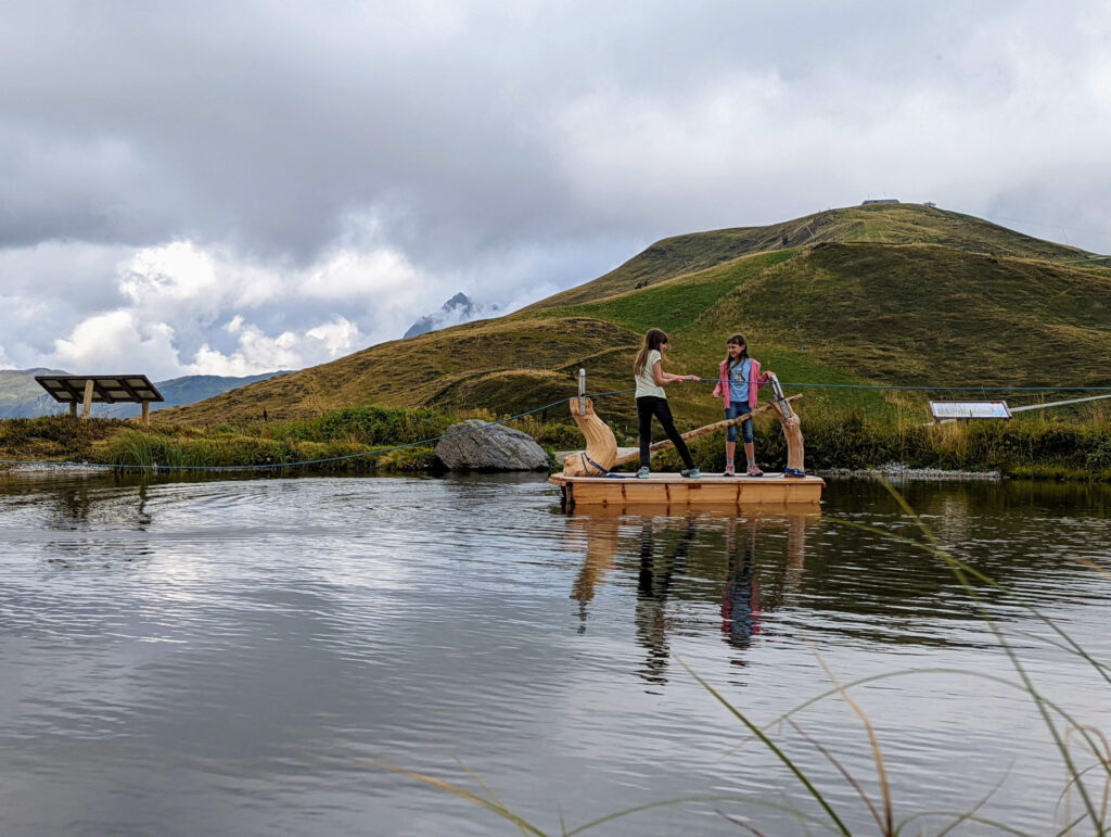 Zwei Kinder stehen auf einem Holzfloß auf einem Speichersee am Resterkogel, ein Abenteuer im Familienurlaub in den Hohen Tauern.