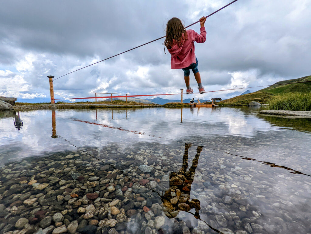 Ein Kind balanciert auf einem Seil über dem flachen Wasser eines Speichersees am Resterkogel, Spiel und Spaß im Familienurlaub.