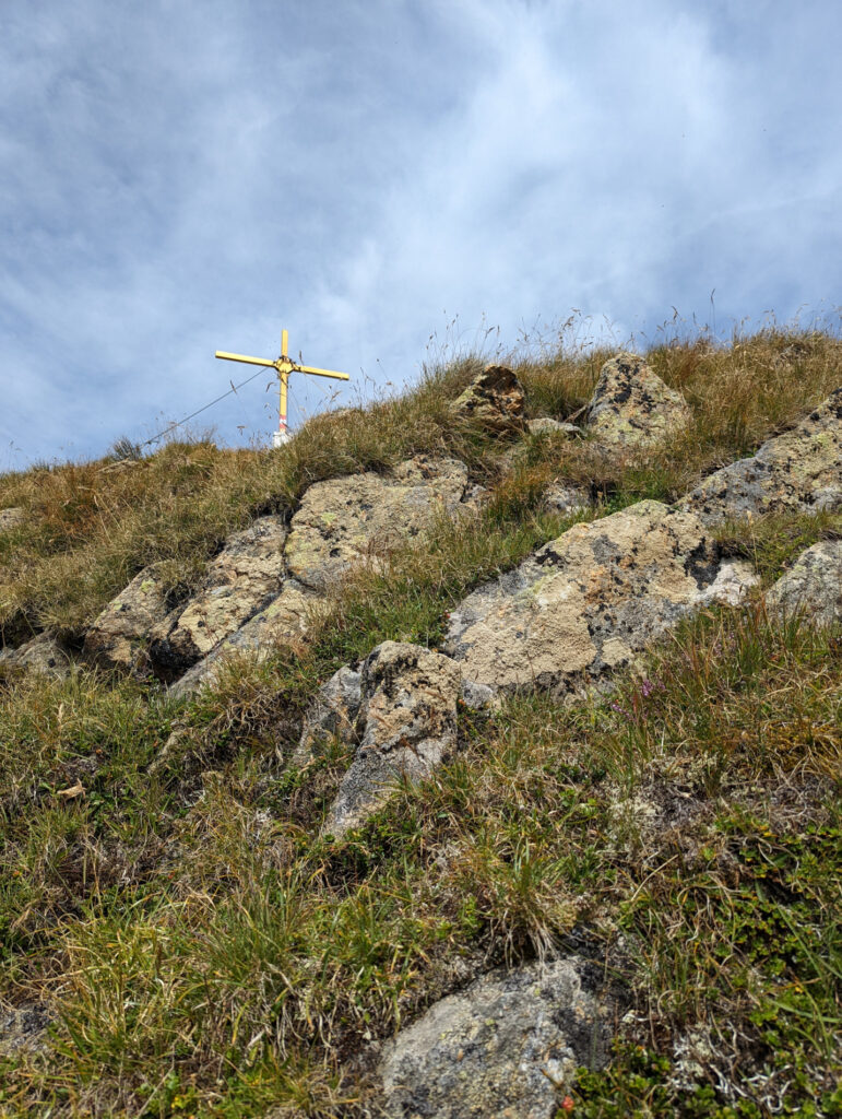 Goldenes Gipfelkreuz auf dem Zwölferkogel, umgeben von felsigem und grasigem Gelände, ein Höhepunkt im Wanderurlaub im Nationalpark Hohe Tauern.