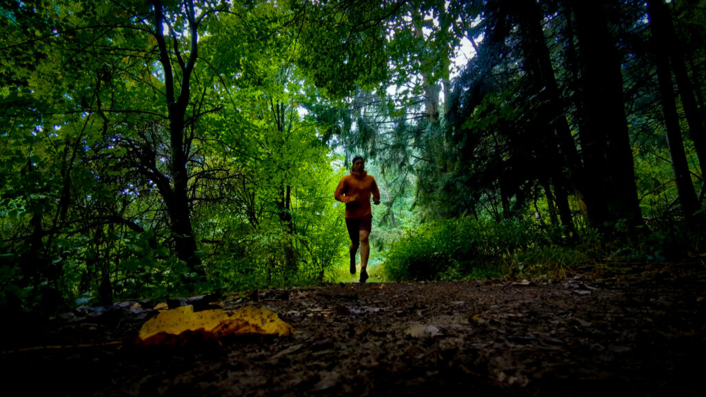 Ein Läufer in einer orangefarbenen Montane Jacke rennt auf einem schmalen Waldweg auf den Betrachter zu. Der Weg ist von dichtem Grün und Bäumen umgeben.