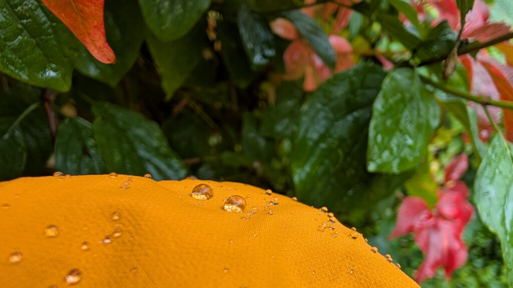 Nahaufnahme des orangefarbenen Stoffes einer Jacke, auf dem viele kleine Wassertropfen liegen und deutlich sichtbar abperlen. Im unscharfen Hintergrund sind grüne und rote Blätter zu sehen.