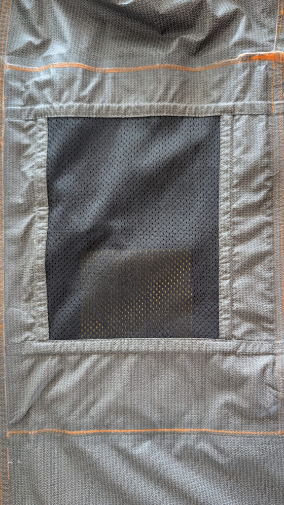 Nahaufnahme der Innenseite einer grauen Jacke mit einer rechteckigen, schwarzen Meshtasche, umgeben von einem grau-orangen Muster.