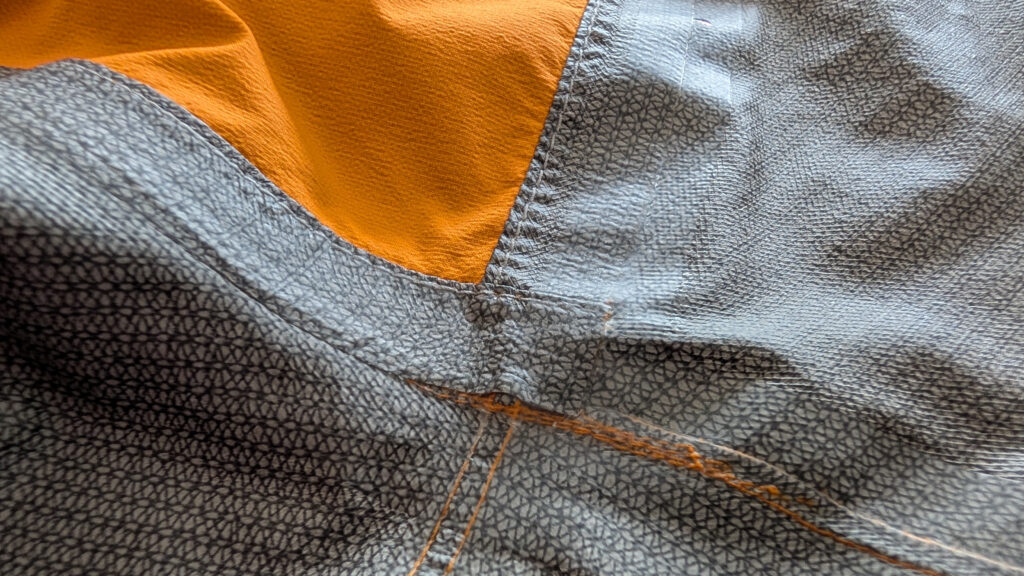 Nahaufnahme von verschiedenen Stoffteilen einer Jacke: ein orangefarbenes Stück, ein graues mit feiner Struktur und eine genähte Naht in der Farbe Orange.
