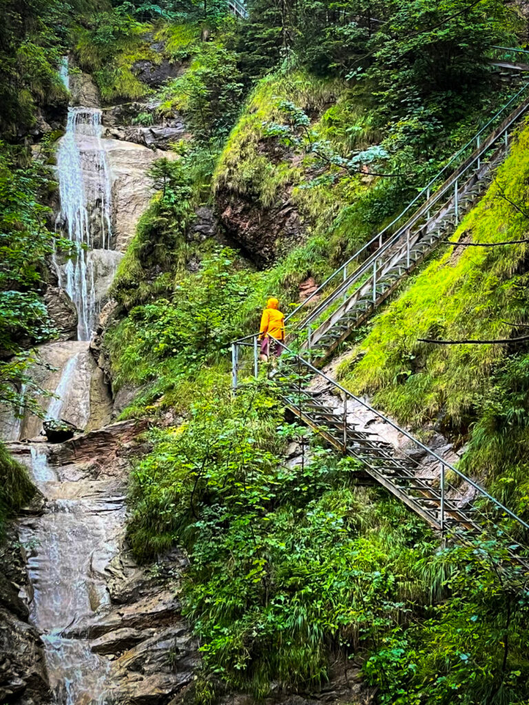 Eine Person in einer leuchtend gelben Montane Jacke steigt eine steile, metallene Treppe neben einem Wasserfall und einem dicht bewachsenen Berghang hinauf. Die Jacke hebt sich stark von der grünen Umgebung ab.