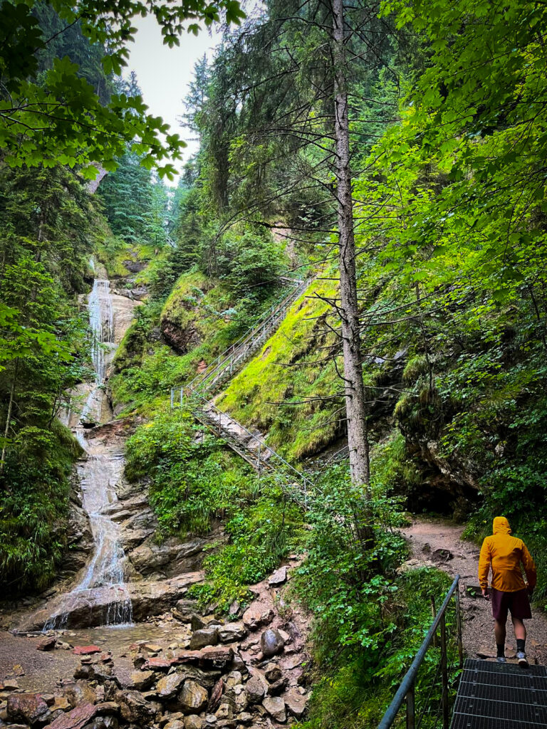 Eine Person von hinten, die auf einem metallenen Steg steht und eine gelbe Jacke sowie rote Shorts trägt. Der Blick geht nach vorne auf einen Wasserfall und einen steilen, bewaldeten Hang mit Treppen.