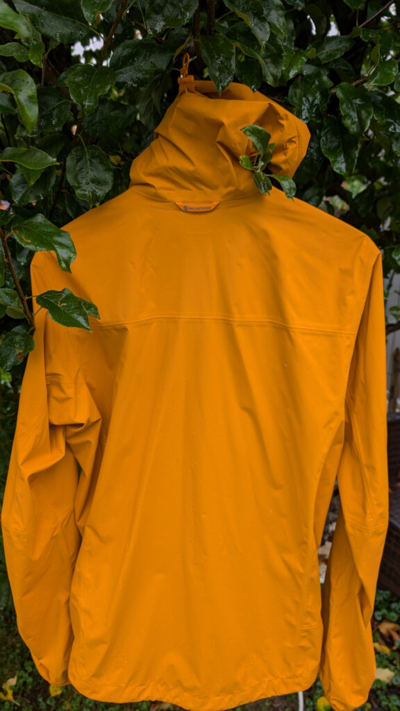 Die Rückseite einer orangefarbenen Jacke mit Kapuze hängt draußen an einem Baum. Blätter mit Wassertropfen sind im oberen Teil des Bildes sichtbar.