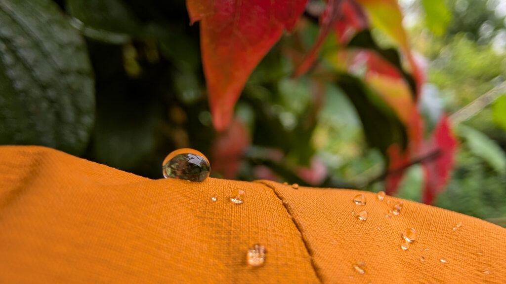 Extrem-Nahaufnahme eines großen Wassertropfens auf dem orangefarbenen Stoff einer Jacke. Im Hintergrund sind unscharfe grüne und rötliche Blätter zu sehen.