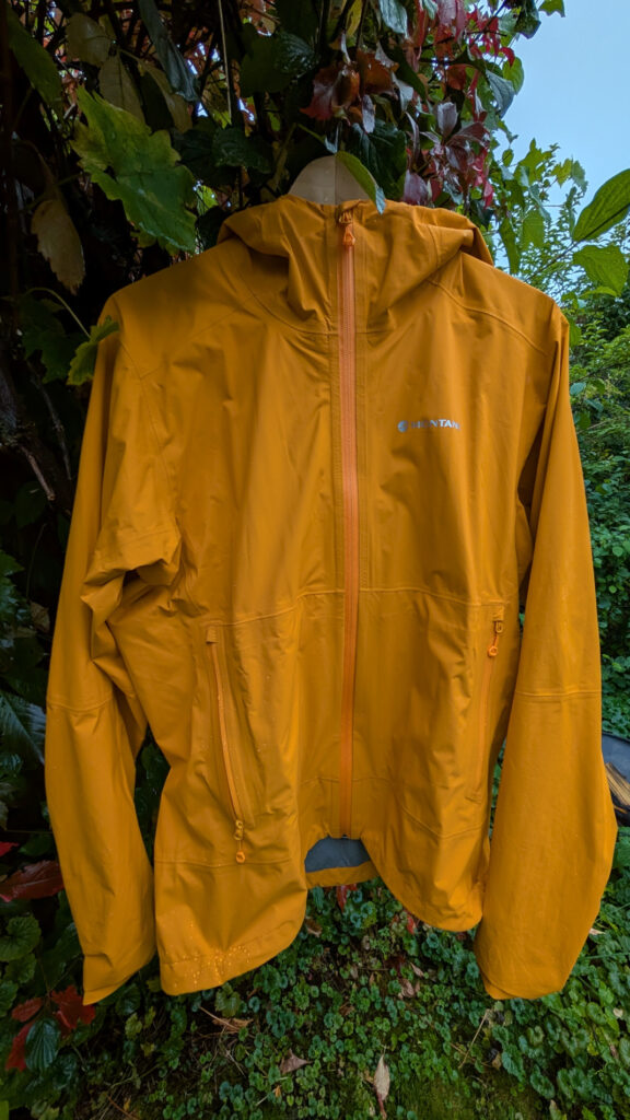 Die Vorderseite einer orangefarbenen Montane Jacke mit Reißverschluss und Bauchtaschen hängt draußen an einem Ast. Im Hintergrund sind grüne und rote Blätter sichtbar.